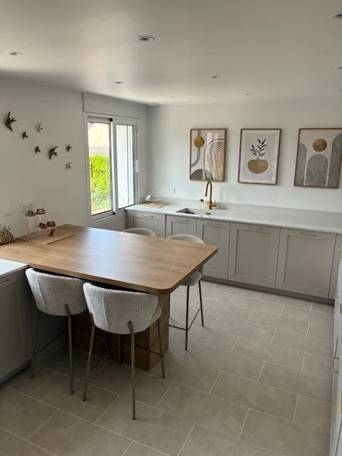 Cuisine lumineuse et moderne avec plan de travail central en bois, îlot-bar et tabourets pastel; rangements gris clair et décor mural végétal. intégration naturelle de “comment moderniser une cuisine”.